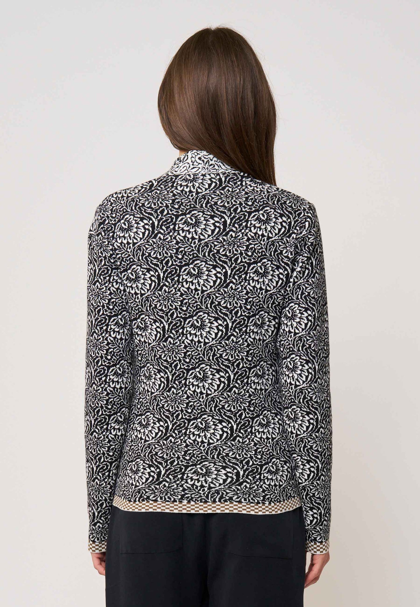 Frau trägt Strickjacke, Modell Jolie, in Schwarz mit weißem floralem Muster, Rückansicht, leichter Saumabschluss