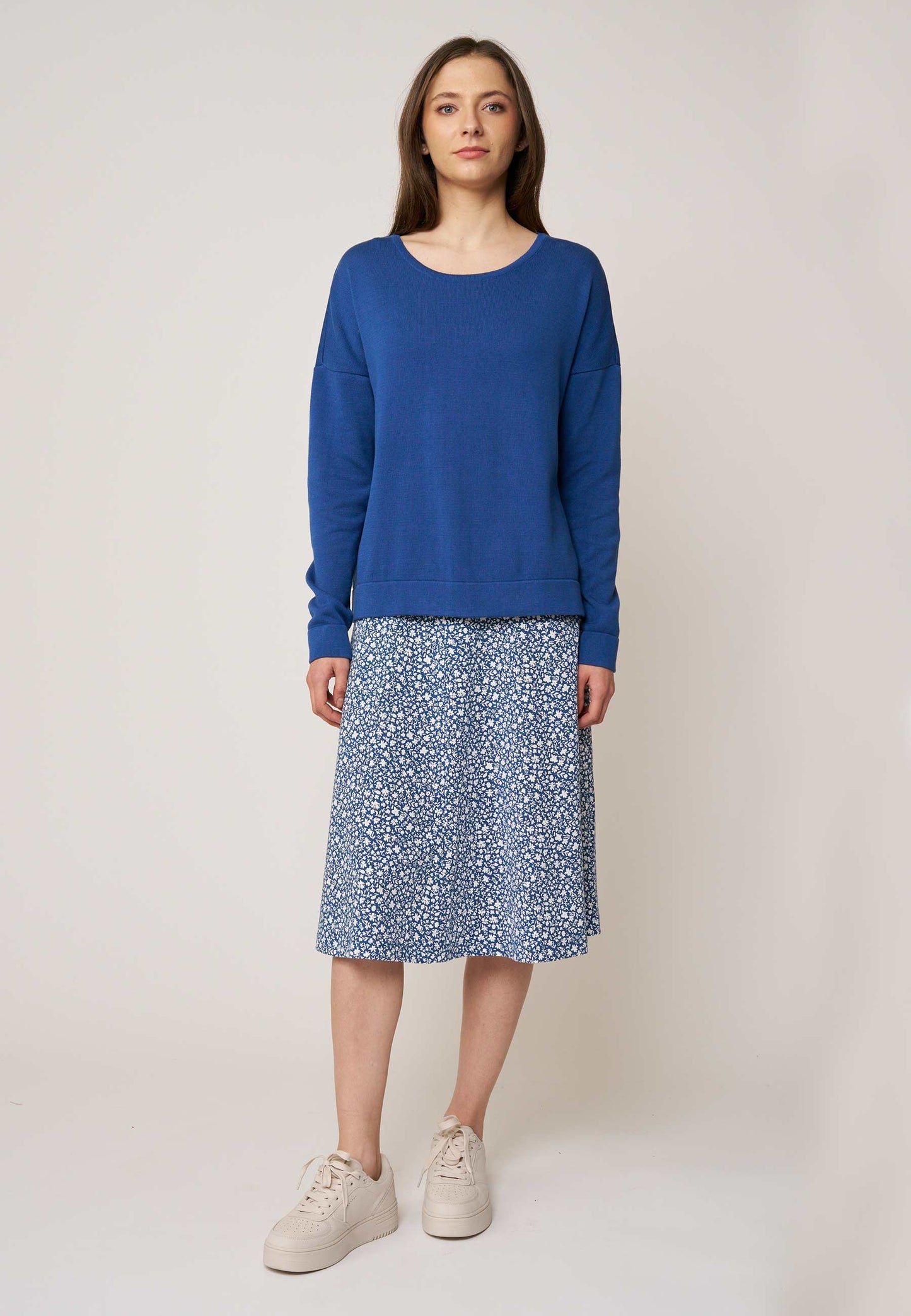 Frau trägt schlichtes, langärmeliges blaues Oberteil mit rundem Ausschnitt und geblümten Midirock in Blau und Weiß