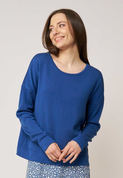 Frau mit langem braunem Haar trägt locker sitzendes, blaues Oberteil mit Rundhalsausschnitt und floral gemustertem Rock