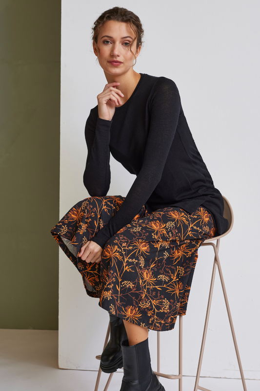 Frau sitzt auf Stuhl trägt schwarzen Pullover und knielangen Rock, Modell Ophelia mit orangefarbenem Blumenmuster